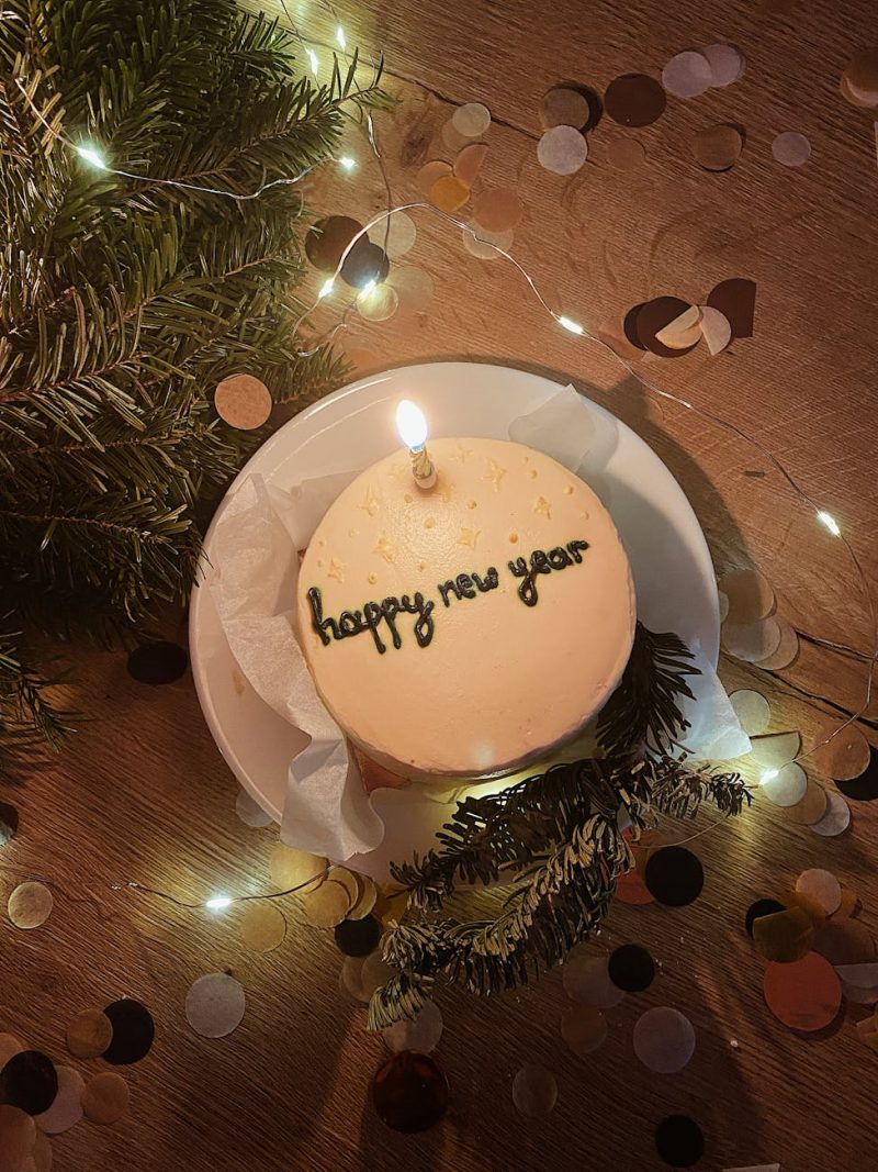 cake with the inscription happy new year with a burning candle on the background of lights and confetti